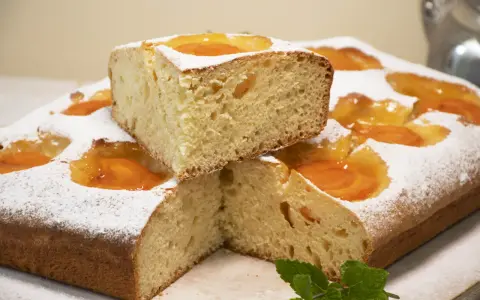 Deliciosa coca de albaricoque, cubierta de azúcar glas y decorada con trozos de fruta. Un trozo apilado revela su esponjosa miga.