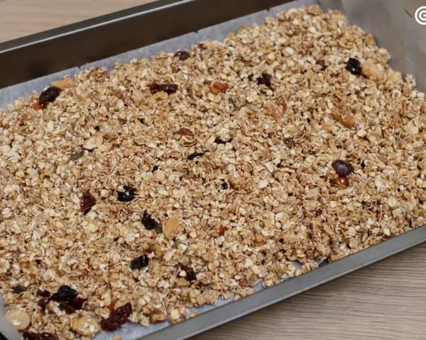 Granola de avena casera lista para hornear, con frutos secos y pasas, extendida en una bandeja.