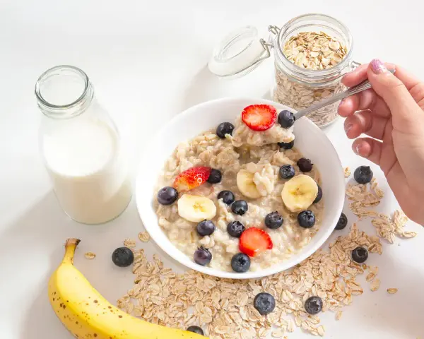 Un tazón de avena con fresas, arándanos y plátano. Un frasco de avena y una botella de leche sugieren cómo se hacen los copos de avena.