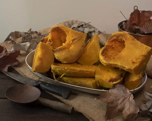 Trozos de calabaza asada, tierna y dorada, servidos en un plato blanco sobre arpillera. Acompañada de utensilios rústicos y hojas secas, evoca el sabor del otoño.