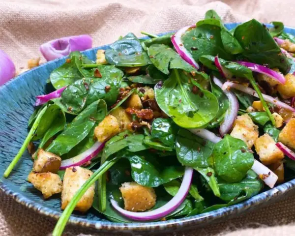Ensalada de espinacas frescas con crutones, cebolla morada y aderezo en un plato azul.