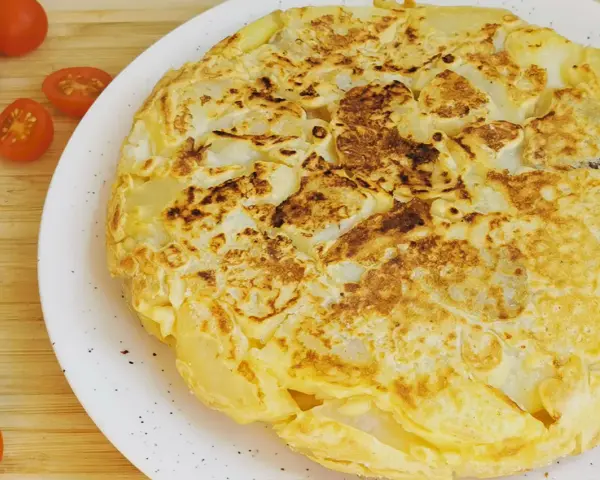 Tortilla vegana dorada y esponjosa, con trozos de patata visibles, servida en plato blanco. Tomates cherry al lado.