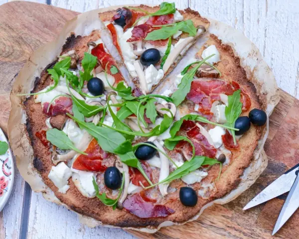Pizza de quinoa con jamón serrano, aceitunas negras, champiñones y rúcula fresca. Una delicia saludable.