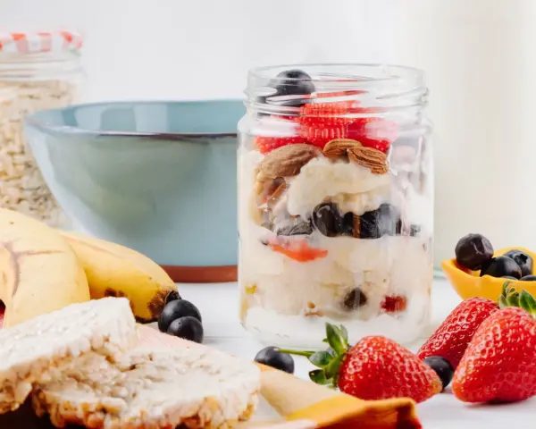 Deliciosa avena nocturna en un frasco con fresas, arándanos y almendras. Plátanos y galletas de arroz al lado.
