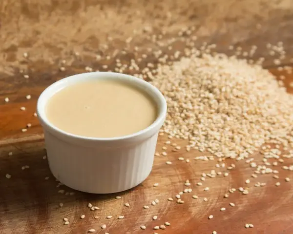 Un cuenco blanco con cremosa salsa de tahini junto a una pila de semillas de sésamo sobre una tabla de madera.