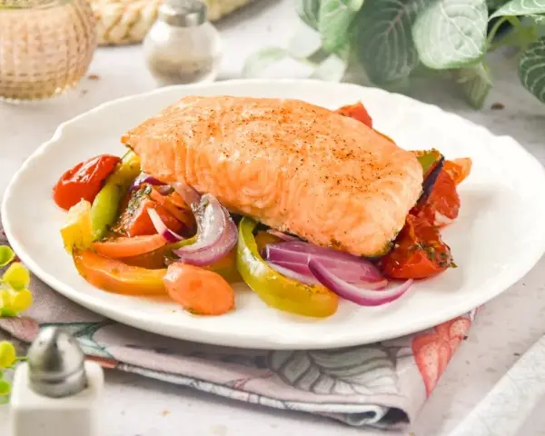 Un jugoso salmón al horno reposa sobre una cama de verduras asadas: tomates, pimientos y cebolla morada.
