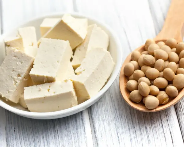 Tofu en cubos en un plato blanco junto a una cuchara de madera llena de semillas de soja. ¿A que sabe el tofu? Neutro, absorbe sabores.