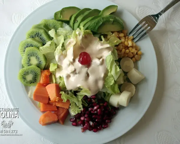 Ensalada tropical con aguacate, kiwi, papaya, palmitos, maíz y granada, cubierta de aderezo cremoso y una cereza.