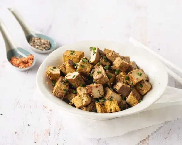 Tazón lleno de cubos dorados de tofu marinado, espolvoreados con cebollín y semillas.