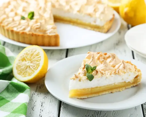 Un delicioso pastel de limón con merengue dorado, acompañado de limones frescos y una servilleta a cuadros verdes. Los ingredientes del lemon pie lucen perfectos.