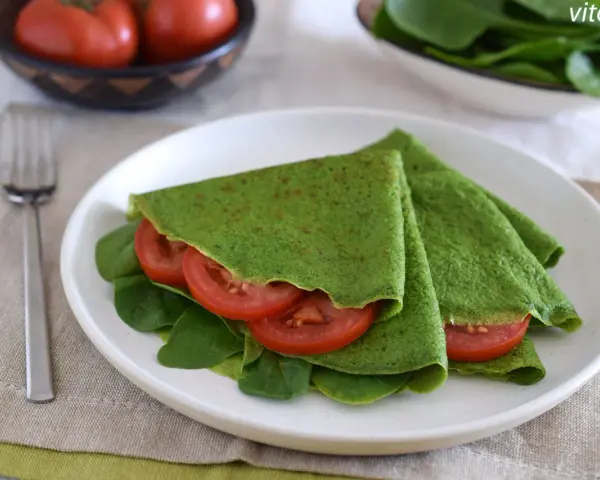 Deliciosos crepes de espinacas rellenos de rodajas de tomate fresco y hojas de espinaca, servidos en un plato blanco.