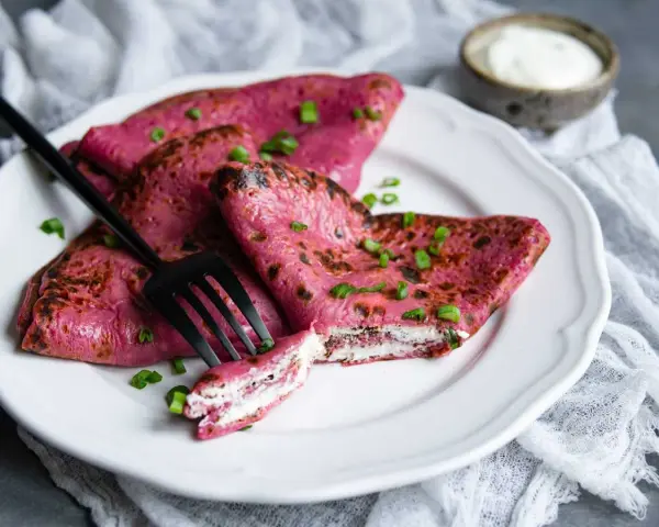 Crepes rosados ​​rellenos de queso crema, perfectos para recetas con remolacha cruda. Decorados con cebollín y acompañados de una porción de crema agria.