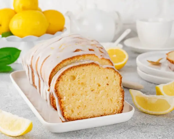 Un delicioso budín de limón glaseado, con rodajas de limón fresco y una taza de té al fondo.