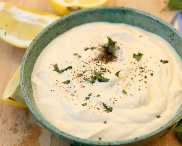 Un cremoso tazón de mayonesa de tofu, adornado con perejil fresco y pimienta negra, junto a rodajas de limón.