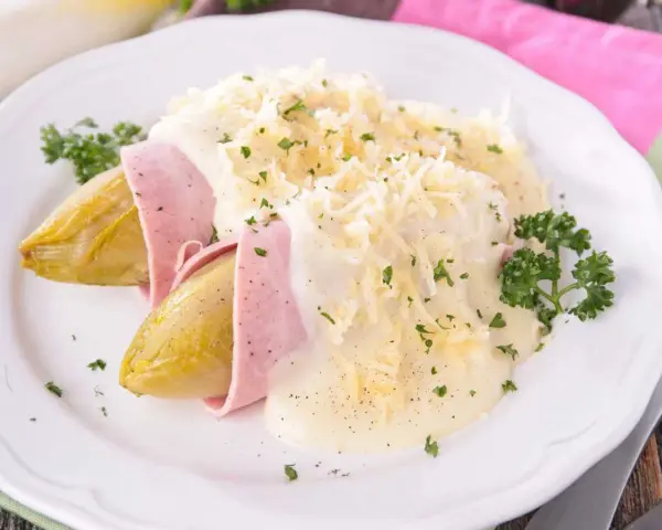 Endivias al horno envueltas en jamón, cubiertas con una cremosa salsa bechamel y queso rallado, adornadas con perejil fresco.