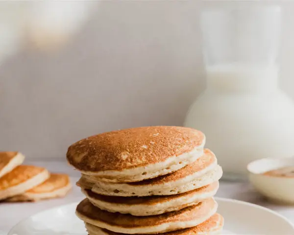 Una pila de esponjosos **pancakes de avena** dorados, con un jarro de leche y un tazón de canela al fondo.