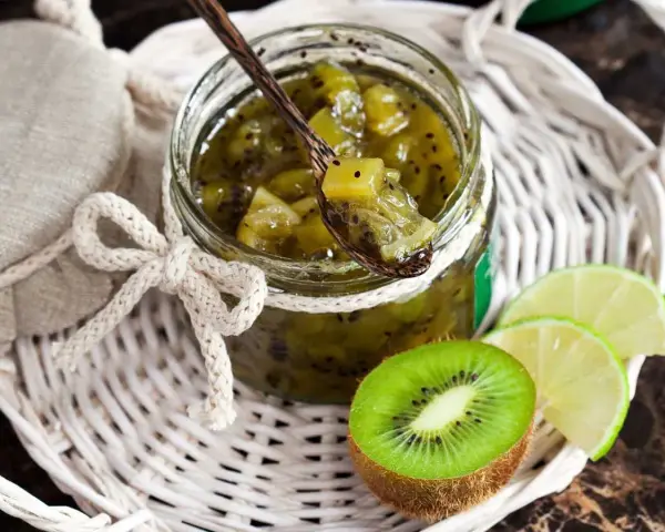 Tarro de mermelada de kiwi casera, con una cuchara de madera llena de la dulce conserva. Rodajas de kiwi y lima acompañan la presentación.