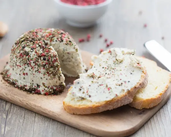 Queso vegano receta: un bloque de queso cremoso cubierto de pimienta y hierbas, servido con rebanadas de pan.