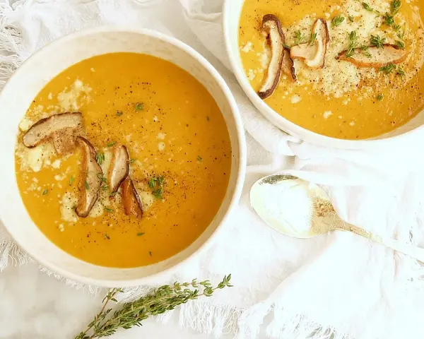 Dos tazones de cremosa sopa de calabaza, adornados con champiñones y queso rallado, listos para ser disfrutados.