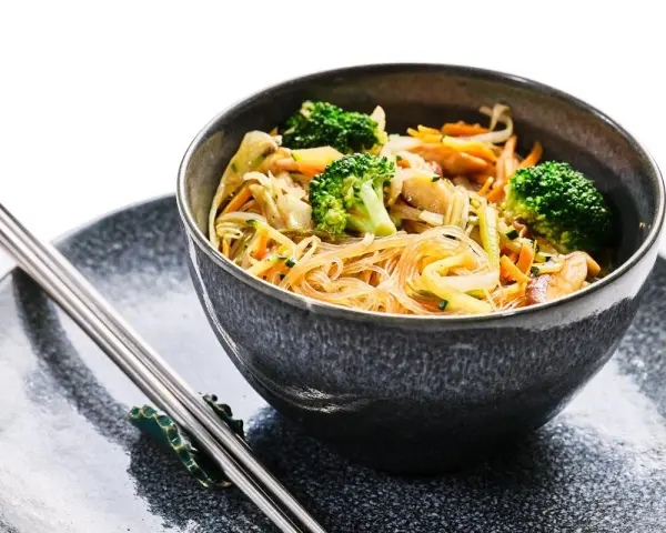 Deliciosa receta con noodles de arroz, brócoli, zanahoria y otros vegetales frescos en un tazón gris, acompañado de palillos.