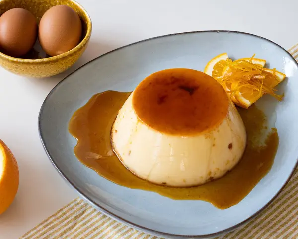 Delicioso flan de naranja con caramelo líquido, decorado con rodajas y ralladura de naranja. Huevos en un cuenco al fondo.