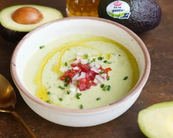 Un cremoso gazpacho de aguacate, adornado con tomate picado, cebolla y cebollino, con un chorrito de aceite de oliva.