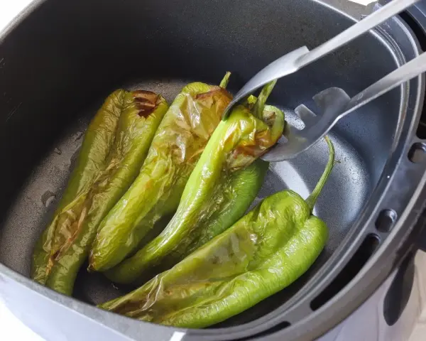 Pimientos verdes asándose en una freidora de aire. Unas pinzas metálicas sujetan uno de los pimientos, mostrando su piel ligeramente chamuscada.