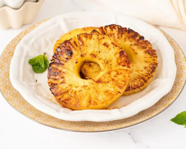 Dos rodajas de piña a la plancha, doradas y caramelizadas, servidas en un plato blanco con hojas de menta.