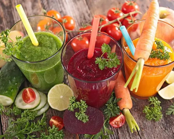 Tres zumos astringentes de colores vibrantes: verde, rojo y naranja, rodeados de ingredientes frescos como zanahorias, remolachas y tomates.