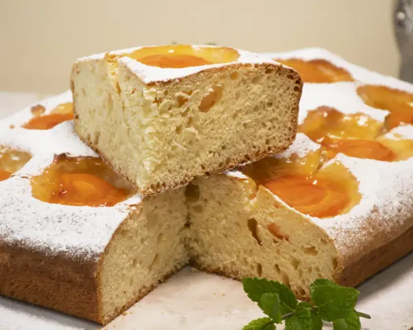 Deliciosa coca de albaricoque, cubierta de azúcar glas y decorada con trozos de fruta. Un trozo apilado revela su esponjosa miga.