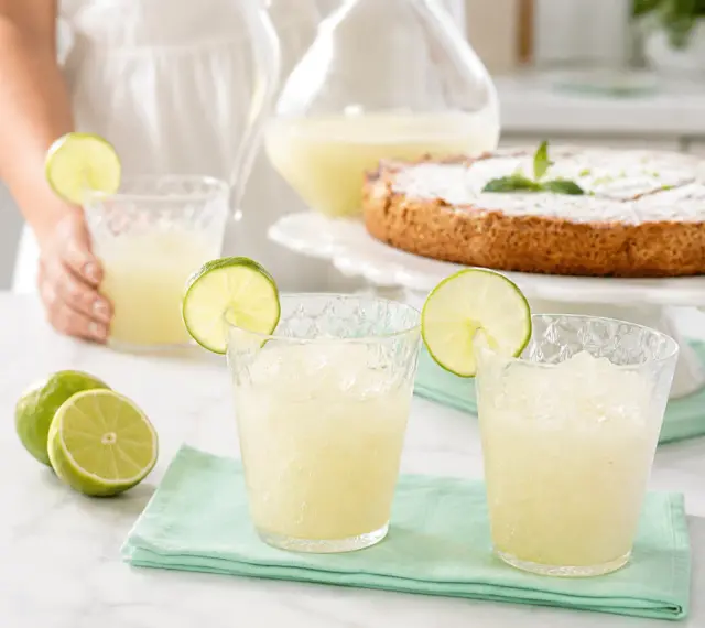 Refrescante granizado de limón servido en vasos con rodajas de lima, junto a limones frescos y un pastel.