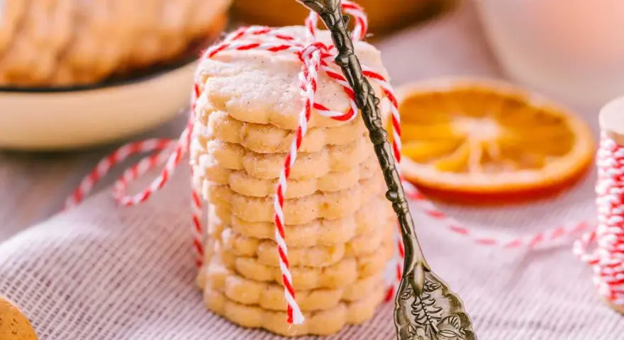 Deliciosas galletas de naranja, apiladas y atadas con hilo rojo y blanco. Una rodaja de naranja y una cuchara antigua completan la escena.