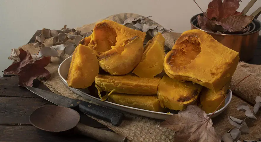Trozos de calabaza asada, tierna y dorada, servidos en un plato blanco sobre arpillera. Acompañada de utensilios rústicos y hojas secas, evoca el sabor del otoño.