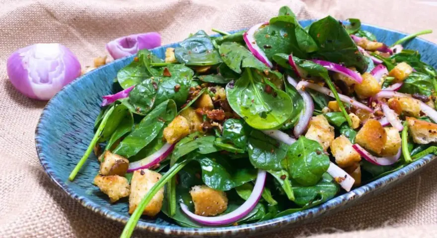 Ensalada de espinacas frescas con crutones, cebolla morada y aderezo en un plato azul.