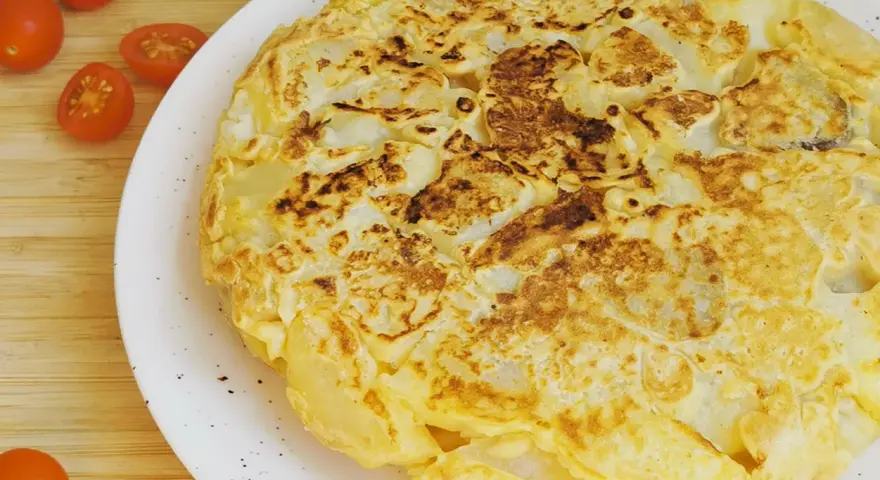 Tortilla vegana dorada y esponjosa, con trozos de patata visibles, servida en plato blanco. Tomates cherry al lado.