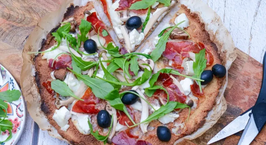 Pizza de quinoa con jamón serrano, aceitunas negras, champiñones y rúcula fresca. Una delicia saludable.