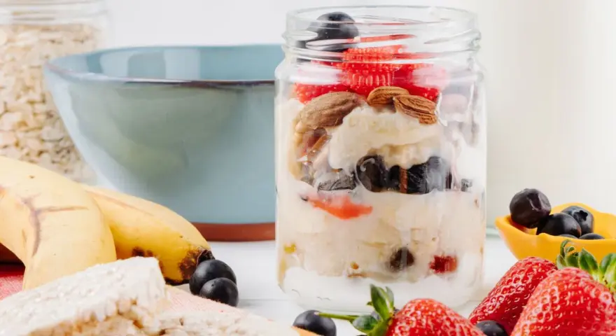 Deliciosa avena nocturna en un frasco con fresas, arándanos y almendras. Plátanos y galletas de arroz al lado.