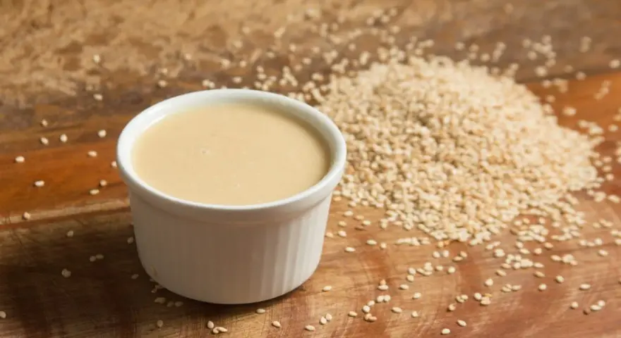 Un cuenco blanco con cremosa salsa de tahini junto a una pila de semillas de sésamo sobre una tabla de madera.
