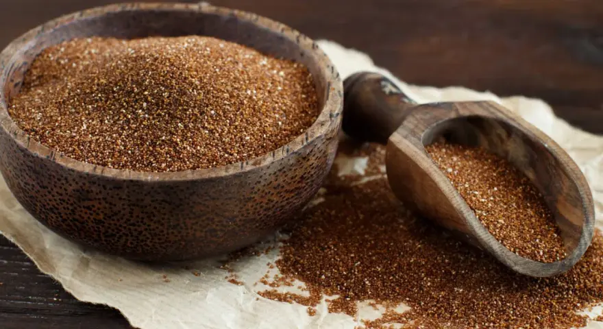 Un cuenco de madera lleno de teff cereal y una pala de madera con más granos.