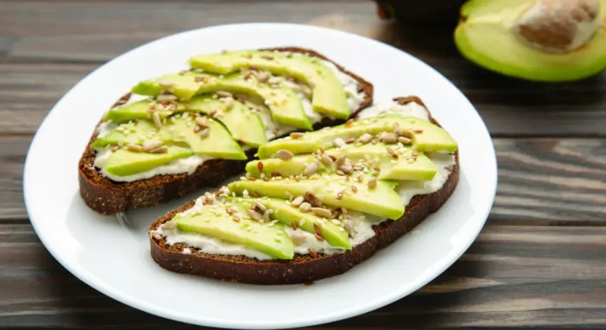 Dos tostadas de pan integral con queso crema, rodajas de aguacate y semillas. Un desayuno saludable y delicioso.