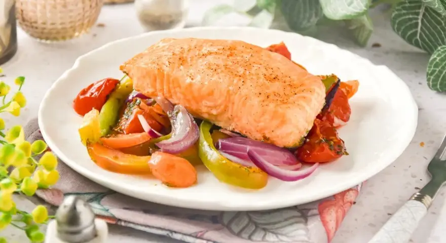 Un jugoso salmón al horno reposa sobre una cama de verduras asadas: tomates, pimientos y cebolla morada.