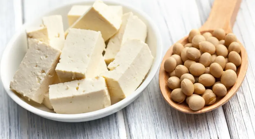 Tofu en cubos en un plato blanco junto a una cuchara de madera llena de semillas de soja. ¿A que sabe el tofu? Neutro, absorbe sabores.