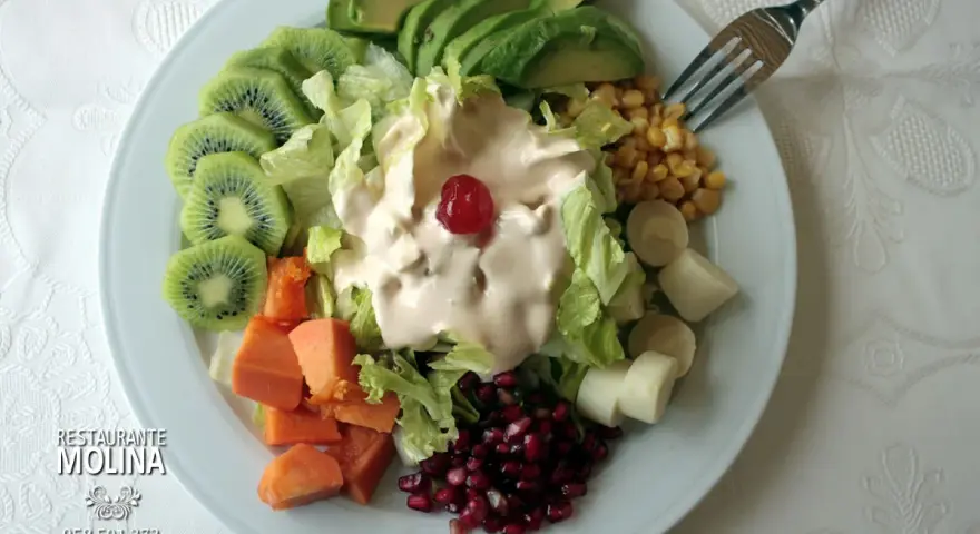 Ensalada tropical con aguacate, kiwi, papaya, palmitos, maíz y granada, cubierta de aderezo cremoso y una cereza.