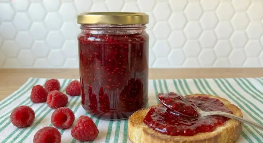 Tarro de mermelada de frambuesa casera, con frambuesas frescas y una tostada untada.