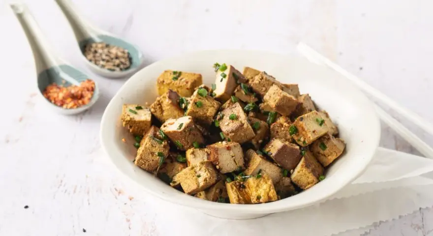 Tazón lleno de cubos dorados de tofu marinado, espolvoreados con cebollín y semillas.