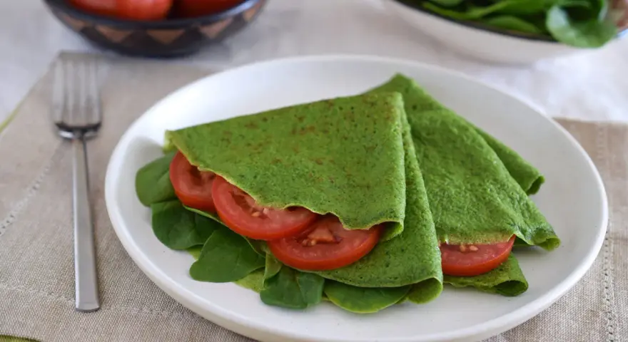Deliciosos crepes de espinacas rellenos de rodajas de tomate fresco y hojas de espinaca, servidos en un plato blanco.