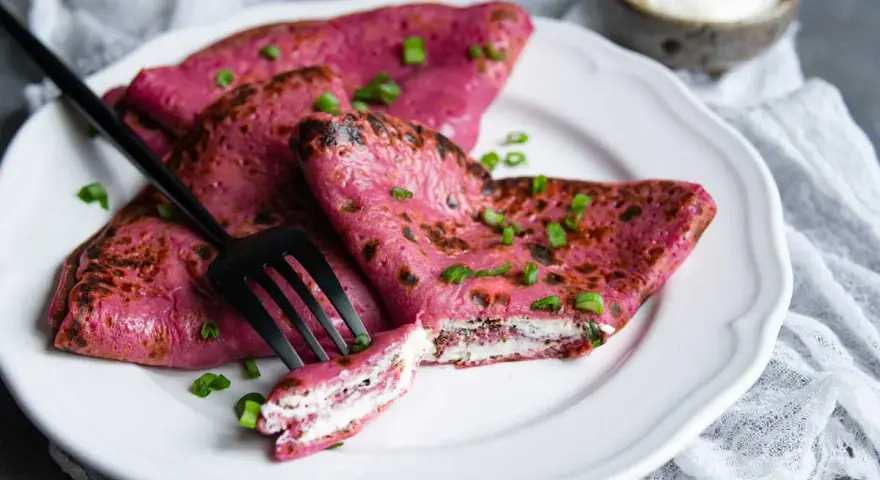Crepes rosados ​​rellenos de queso crema, perfectos para recetas con remolacha cruda. Decorados con cebollín y acompañados de una porción de crema agria.