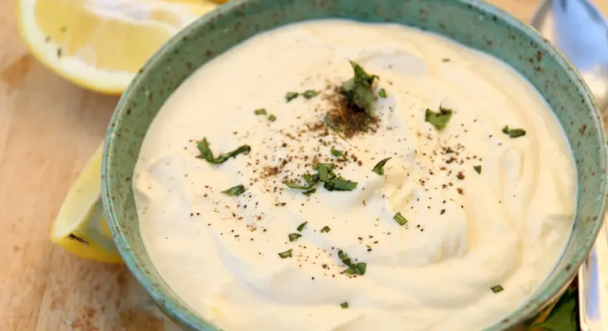 Un cremoso tazón de mayonesa de tofu, adornado con perejil fresco y pimienta negra, junto a rodajas de limón.