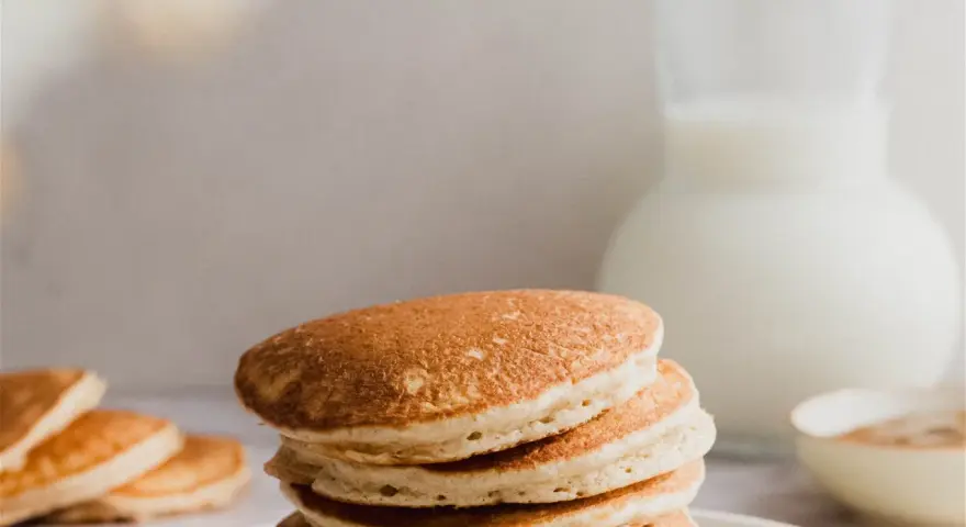 Una pila de esponjosos **pancakes de avena** dorados, con un jarro de leche y un tazón de canela al fondo.
