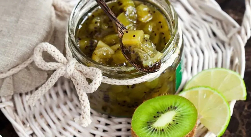 Tarro de mermelada de kiwi casera, con una cuchara de madera llena de la dulce conserva. Rodajas de kiwi y lima acompañan la presentación.